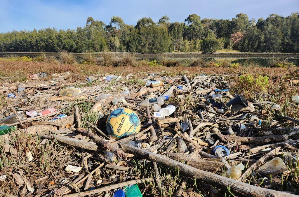 A yellow and blue soccer ball lies amid litter including plastic bottles and cans on a grassy field with trees in the background.