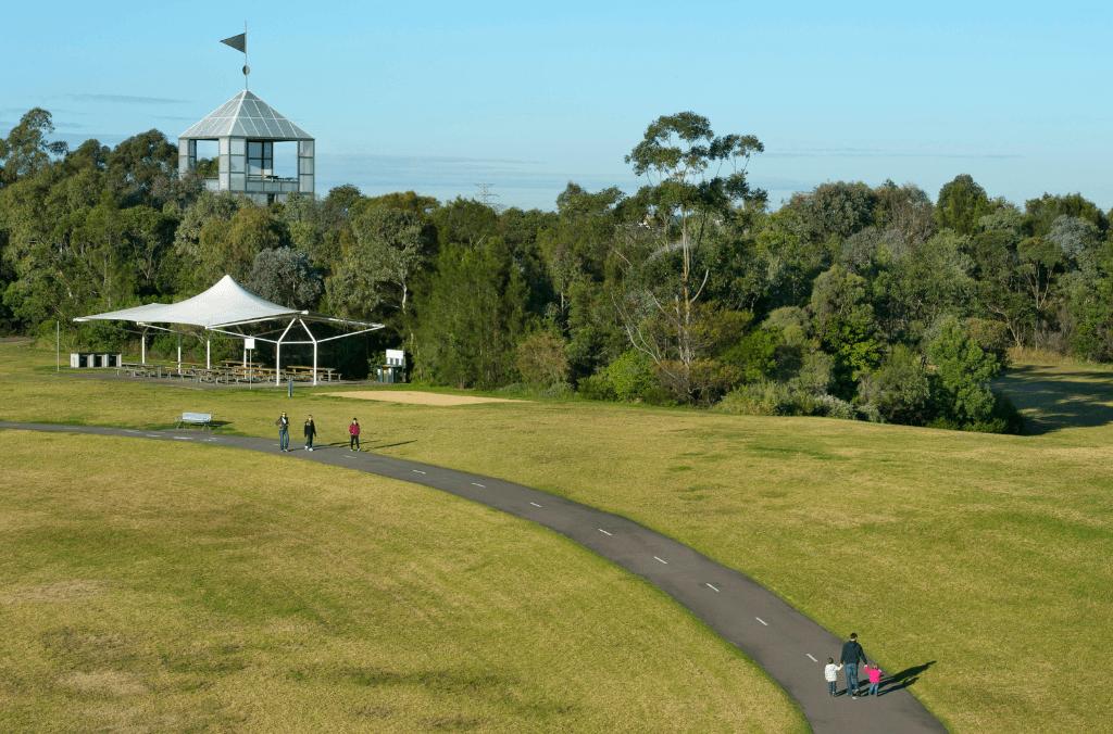 A winding paved path crosses a grassy park where people walk past a shaded picnic shelter, with dense trees and a lookout tower rising in the background under a clear sky.